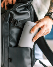 Person slides FELLOW | Carter Move Mug, Matte Black / 16 oz, into backpack pocket, showcasing its portability and secure, spill-proof design for on-the-go coffee enjoyment.