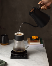 Fellow | Stagg EKG Pro Electric Kettle, Matte Black with Walnut Accents, pours water into a dripper on a marble countertop, embodying precision in coffee brewing.