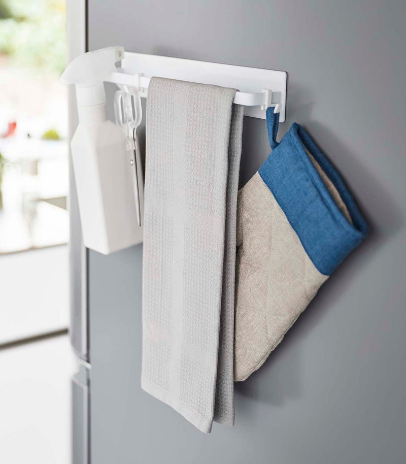 Yamazaki Home's Magnetic Kitchen Towel Hanger, Steel, White, holds a spray bottle, scissors, towel, and oven mitt on a refrigerator side.