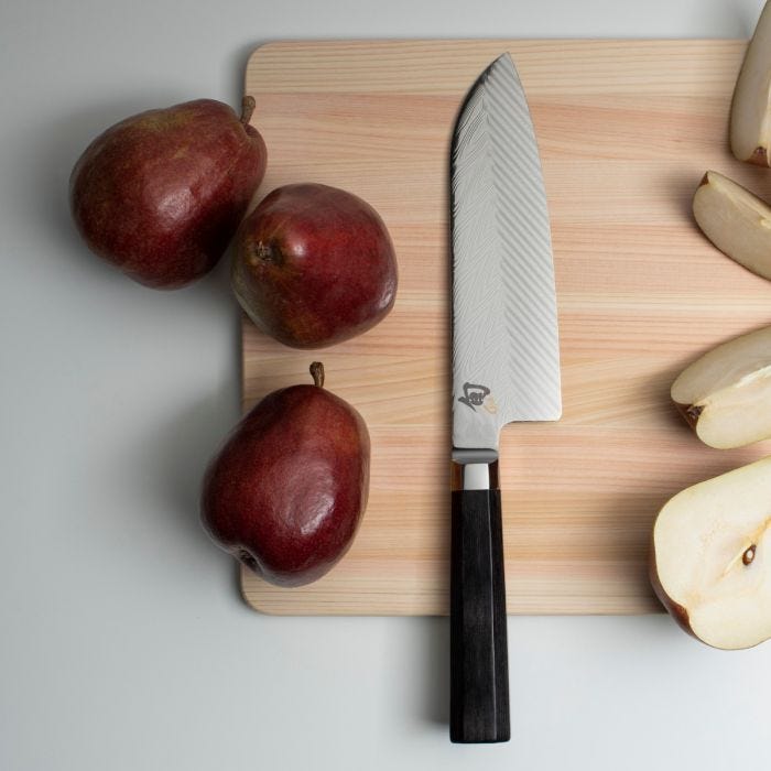 Shun Dual Core Santoku knife on a wooden cutting board with red pears; features a 7-inch patterned blade and dark handle, ideal for versatile kitchen tasks.