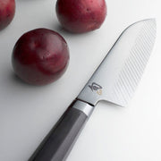 Shun Dual Core Santoku 7-inch knife with Damascus pattern and octagonal handle, displayed on a white surface next to three whole plums.