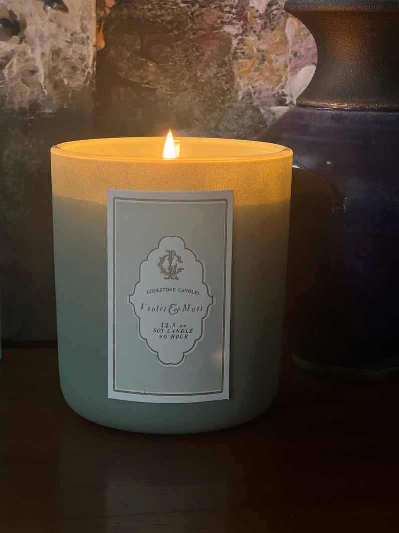 Lodestone Candles | Violet & Moss Candle with a lit flame and labeled jar, displayed on a table, showcasing its elegant design.