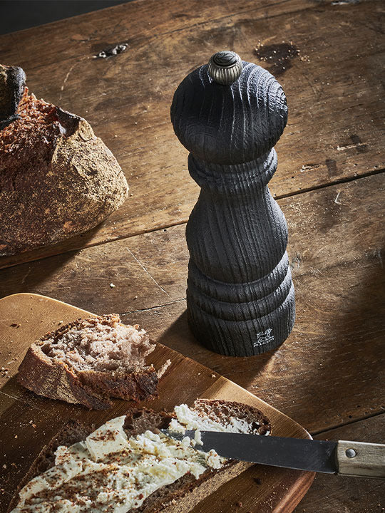 Peugeot | Paris Nature Collection Pepper Mill, Nature Black, on a rustic table with sliced bread and cheese, embodying eco-friendly craftsmanship for culinary excellence.