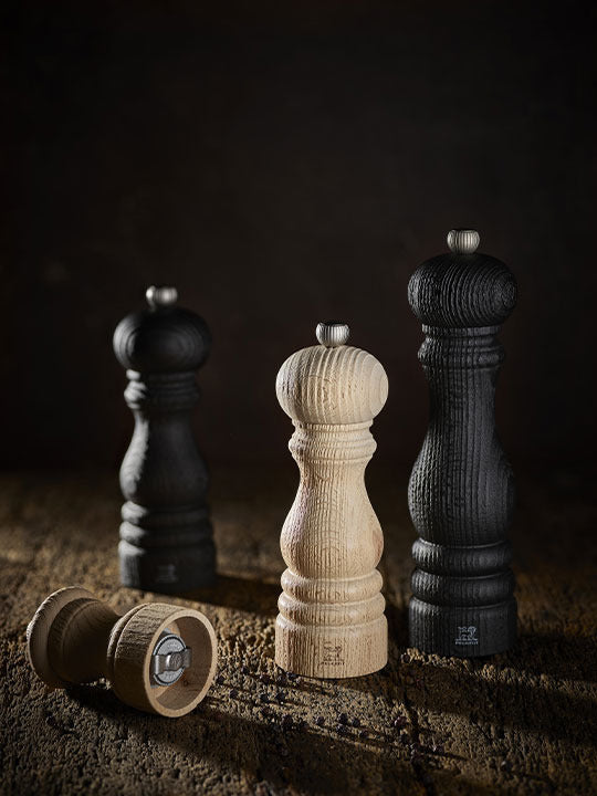 Peugeot | Paris Nature Collection Pepper Mill, Nature Black, alongside other wooden mills, showcases eco-friendly design with a visible grinding mechanism and scattered peppercorns.