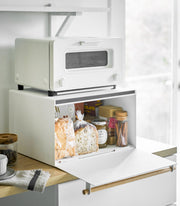 White two-tier Yamazaki Home Simple Steel Bread Box with toaster oven on top, storing bread, jam, and cocoa inside; coffee jar and cup nearby on a wooden counter.