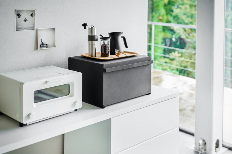 Yamazaki Home's Tower Bread Box in black sits on a sideboard, enhancing kitchen organization alongside a toaster oven and coffee accessories in a minimalist kitchen setting.