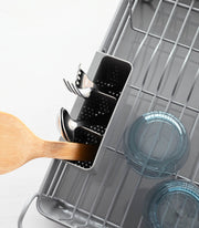 Yamazaki Home | Steel + Wood Dish Rack with Drainer Spout featuring steel frame, wooden handles, and utensil holder, displayed with a dishwasher's cutlery and bowls.