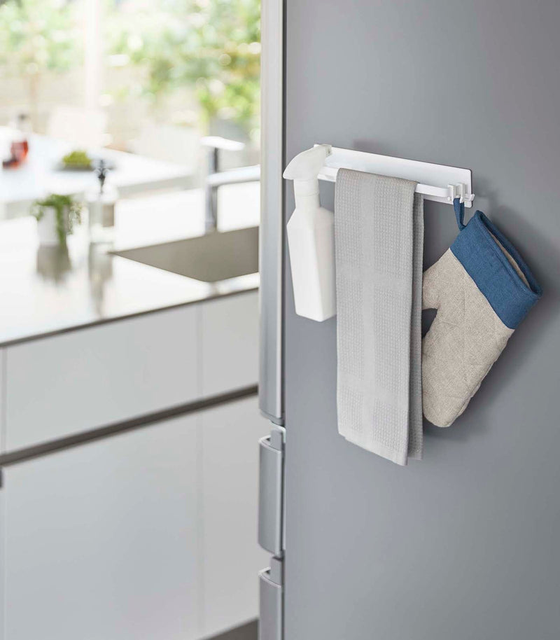 Yamazaki Home | Magnetic Kitchen Towel Hanger, Steel, White on a fridge, holding a spray bottle, gray towel, and blue-trimmed oven mitt in a bright kitchen.