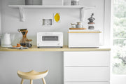 Yamazaki Home Simple Steel Bread Box with wood top, part of a minimalist kitchen setup, offers stylish bread storage alongside a toaster oven and coffee-making essentials.