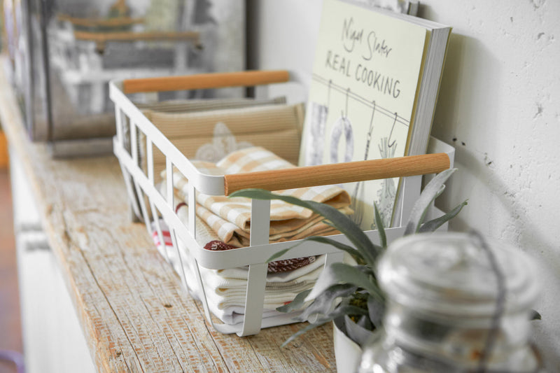 Yamazaki Home | Steel + Wood Storage Baskets, Large, holding folded linens and a cookbook, with a potted plant nearby, creating a cozy kitchen vignette.