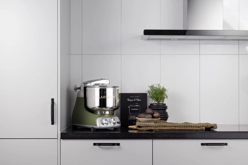 Ankarsrum stand mixer on a black countertop, with stainless-steel bowl, cookbook, herb pot, avocados, and kitchen towel, illustrating versatile kitchen functionality.