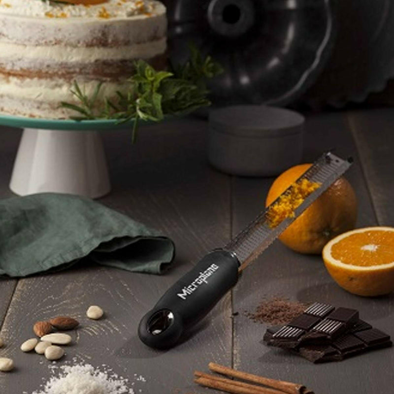 Microplane | Premium Zester, Grater - Classic Blue on a kitchen counter with orange zest, baking ingredients, and a frosted cake in the background.