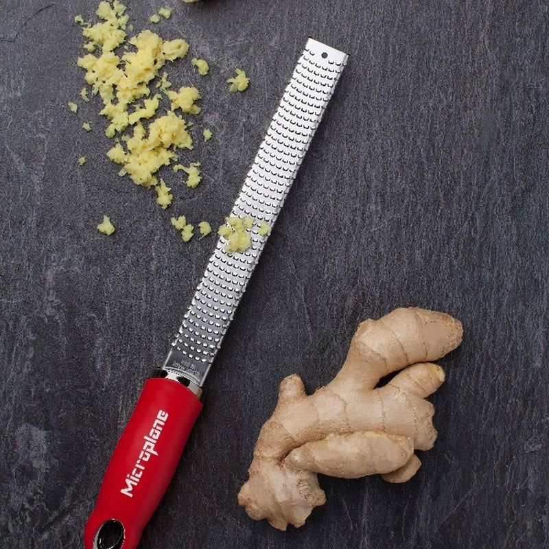 Microplane Premium Zester, Grater on stone surface with fresh ginger and grated shreds, showcasing its efficient stainless-steel blade and ergonomic red handle for culinary precision.