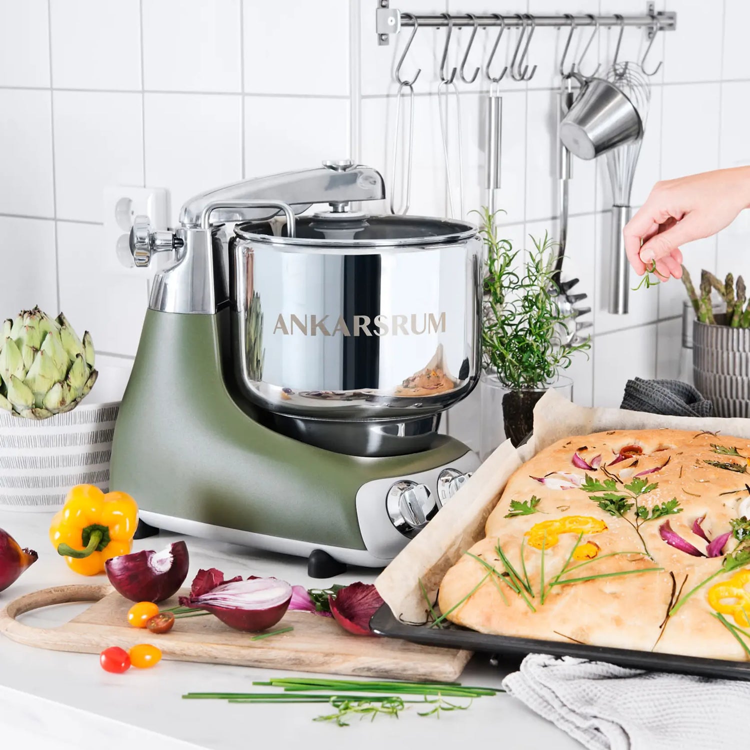 Ankarsrum stand mixer in olive green with steel bowl on a kitchen counter, surrounded by vegetables, fresh focaccia, and a hand adding rosemary.