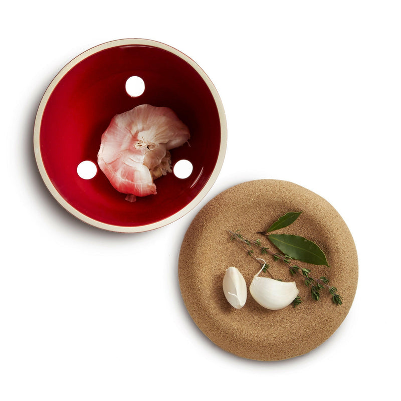 Emile Henry | Garlic Pot - Truffle with vented design, holds a partially peeled garlic head; nearby, a cork trivet displays peeled garlic cloves and fresh herb sprigs.