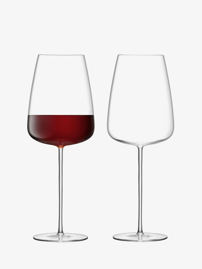 A set of two handmade, mouth-blown red wine glasses with flat-based bowls, alongside a polished, hand-crafted wine carafe with a slanted rim.