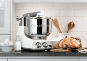 Ankarsrum stand mixer on a countertop with baking essentials, including eggs, milk, and a finished loaf, showcasing its versatility in a kitchen setting.
