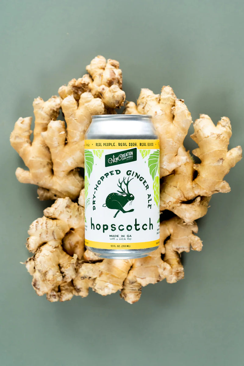 New Creation Soda Works' Krümkake Butter Pecan Cream Soda, a silky, creamy craft soda with caramel, vanilla, and pecan notes, showcased beside ginger root and a green rabbit.