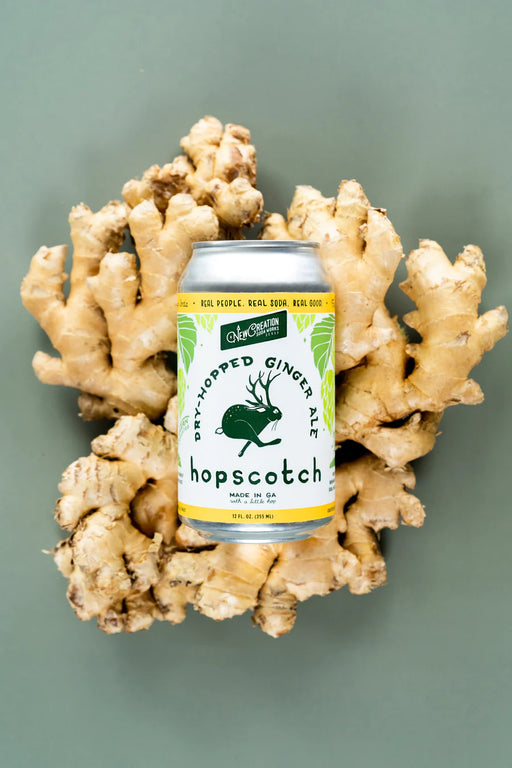 New Creation Soda Works' Krümkake Butter Pecan Cream Soda, a silky, creamy craft soda with caramel, vanilla, and pecan notes, showcased beside ginger root and a green rabbit.
