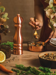Copper Peugeot Paris Chef Pepper Mill on wooden countertop, surrounded by cooking essentials. Hand scoops grain mix from copper pan, embodying elegance and functionality.