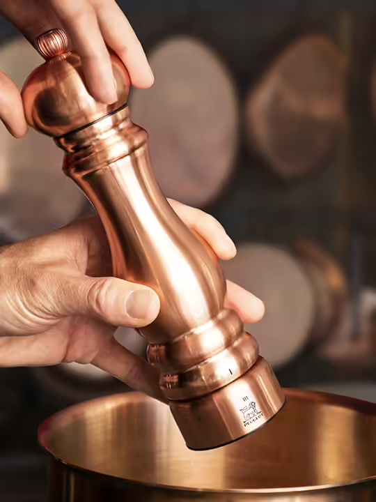 Person using Peugeot | Paris Chef u'Select Copper Pepper Mill over a copper pot, showcasing its ergonomic design and adjustable grind settings in a kitchen environment.