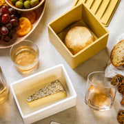 Close-up of the CapaBunga Cheese Vault, a container designed for preserving artisan cheese's freshness, featured on a table with bread and cheese.