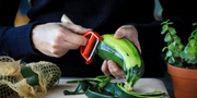 A person peels a zucchini with the Opinel T-Duo Peeler - Green, featuring dual blades, on a wooden board, showcasing its versatility in the kitchen.