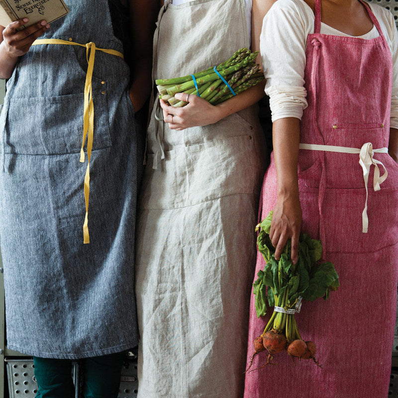 Women in aprons, featuring the Sir|Madam Linen Grand Apron - Faded Black, showcasing four compartments and adjustable neck loop for kitchen use.