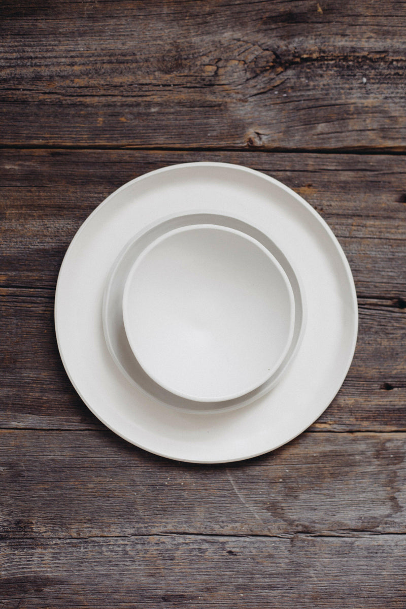 Overhead view of the Looks Like White | Scandi Cereal Bowl nested in a dinner plate on a rustic wooden table, highlighting elegance and simplicity.