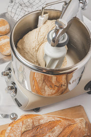 Ankarsrum | Stand Mixers in Store - Cloudy Pink kneads bread dough, with a crusty loaf and flour-dusted rolls nearby on a wooden board.