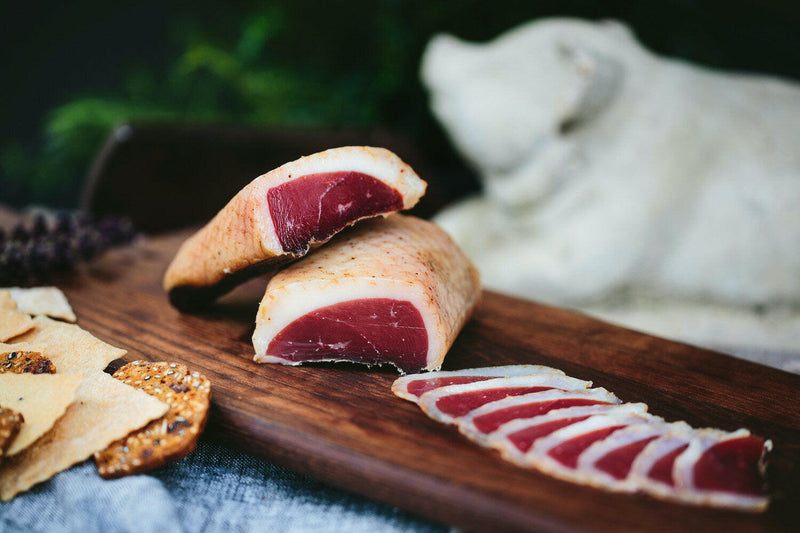 Spotted Trotter | Sliced Charcuterie Meats - Duck Prosciutto displayed on a wooden surface, accompanied by crackers, showcasing its marinated, thin-sliced presentation.