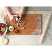 Hands peeling an apple beside the J.K. Adams French Tapered Rolling Pin, showcasing its tapered design for maneuverability, on a flour-dusted marble countertop.