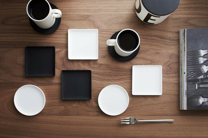 Overhead view of Yamazaki Tower Silicon Coaster set on a wood surface, surrounded by white mugs, dishes, and a fork, showcasing minimalist coffee serving.