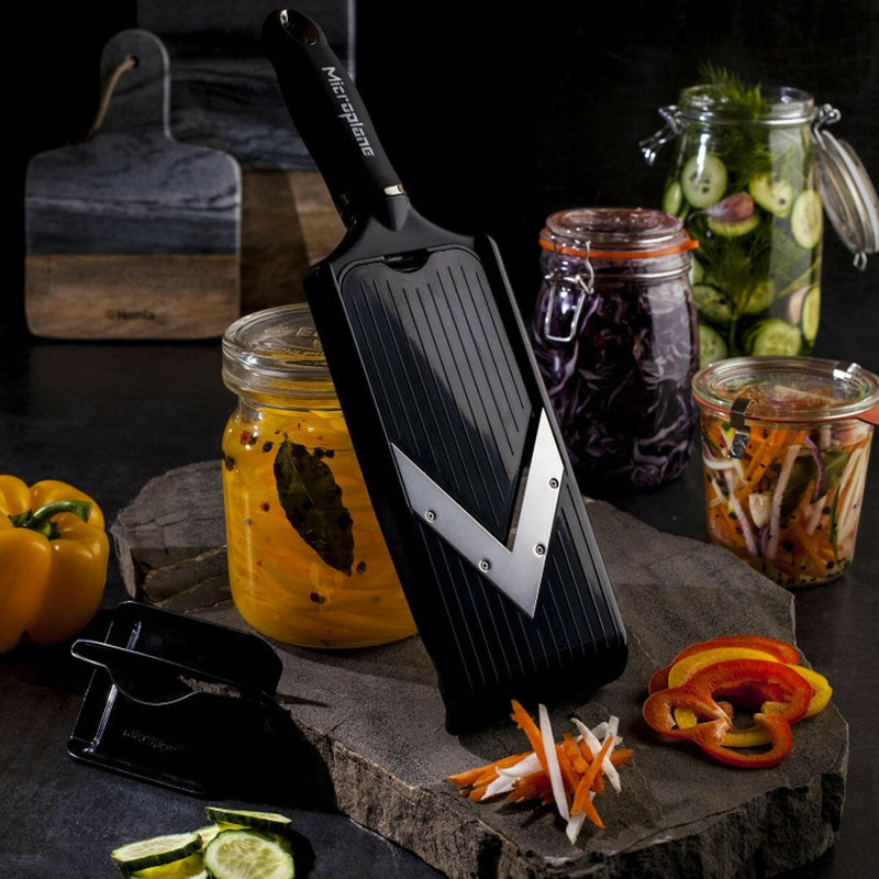 Microplane Adjustable V-Blade Mandoline Slicer amid sliced vegetables and pickled jars, showcasing its precise cutting ability for meal prep.
