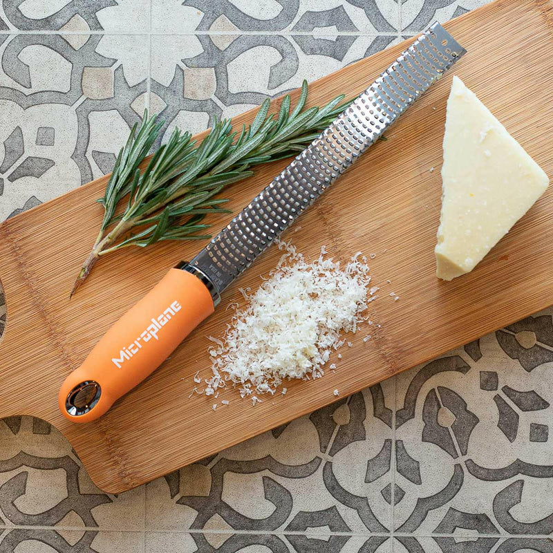 Microplane Premium Zester Grater on a wooden board with grated Parmesan, cheese wedge, and rosemary, showcasing its efficient grating capability for culinary enthusiasts.